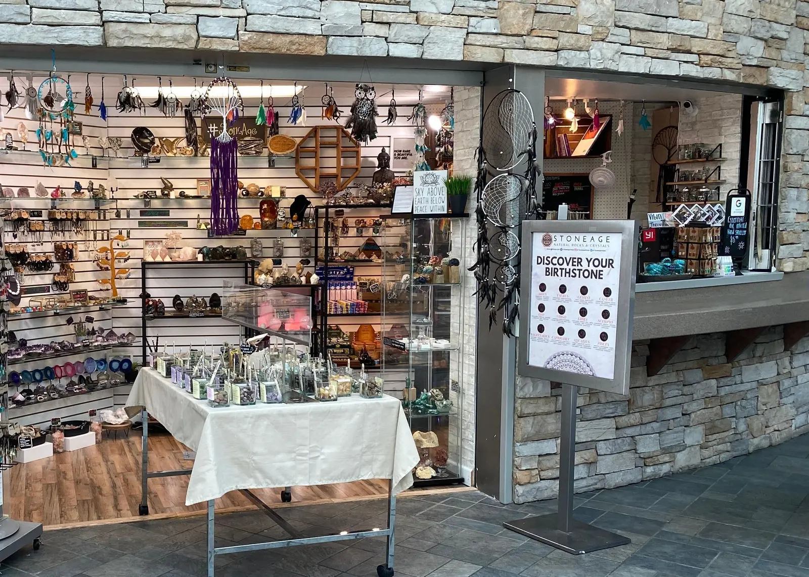 The first Stoneage seen from the mall concourse: a small open unit in a stone wall, its slatwall packed with crystals and dream catchers, a cloth-covered table of tumbled stones inside, and a Stoneage birthstone sign on the walkway