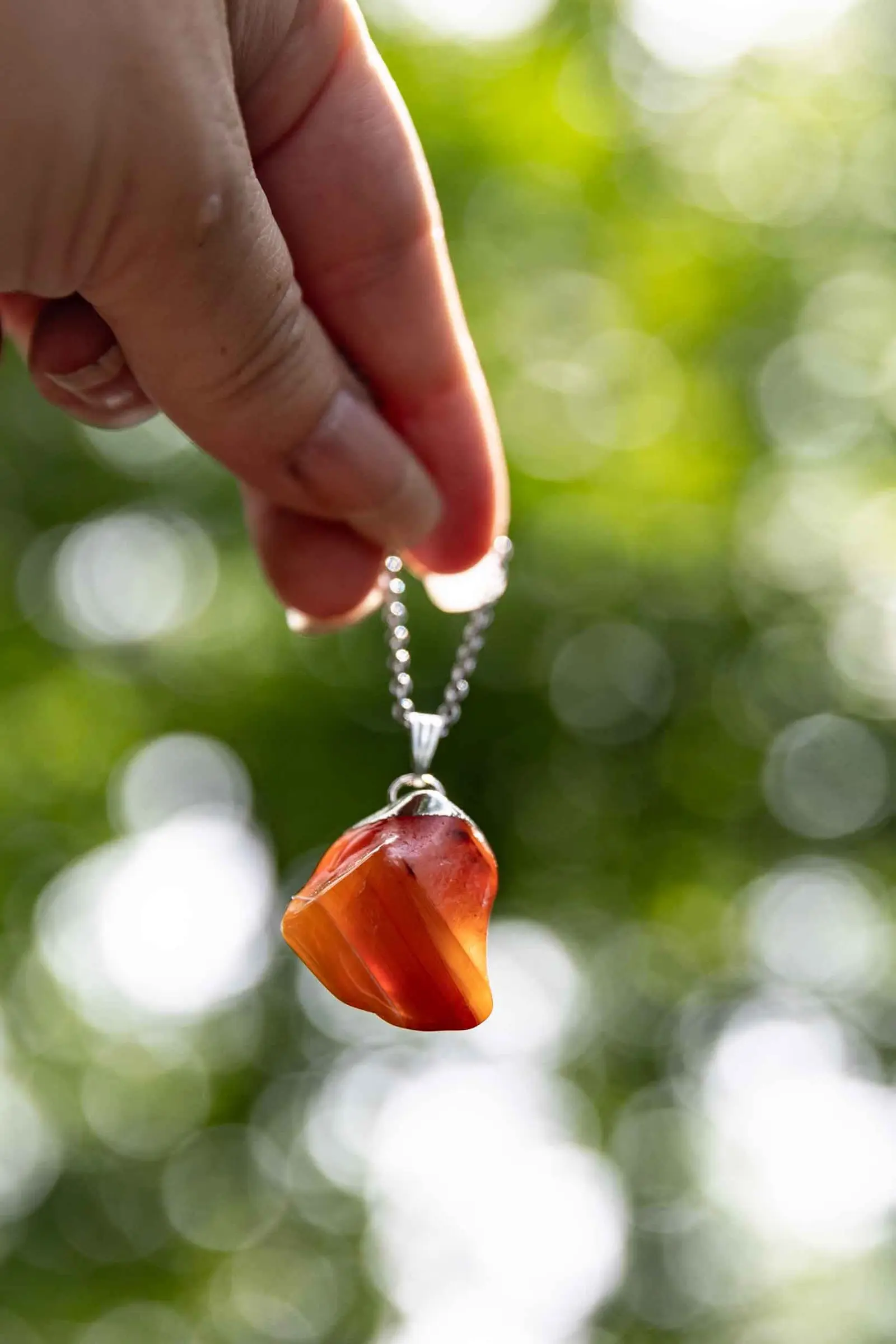 A hand holding a carnelian pendant up to the light on its chain, sunlit woodland behind it