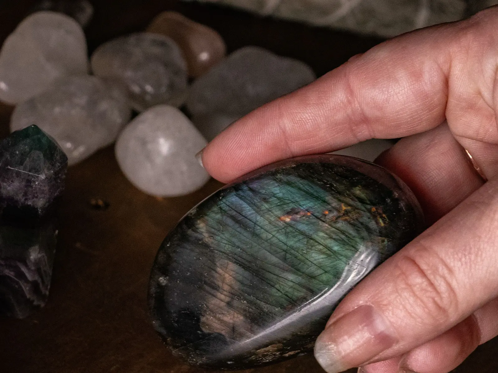 A hand lifting a polished labradorite palm stone from a timber table, tumbled quartz and a bundle of sage behind it