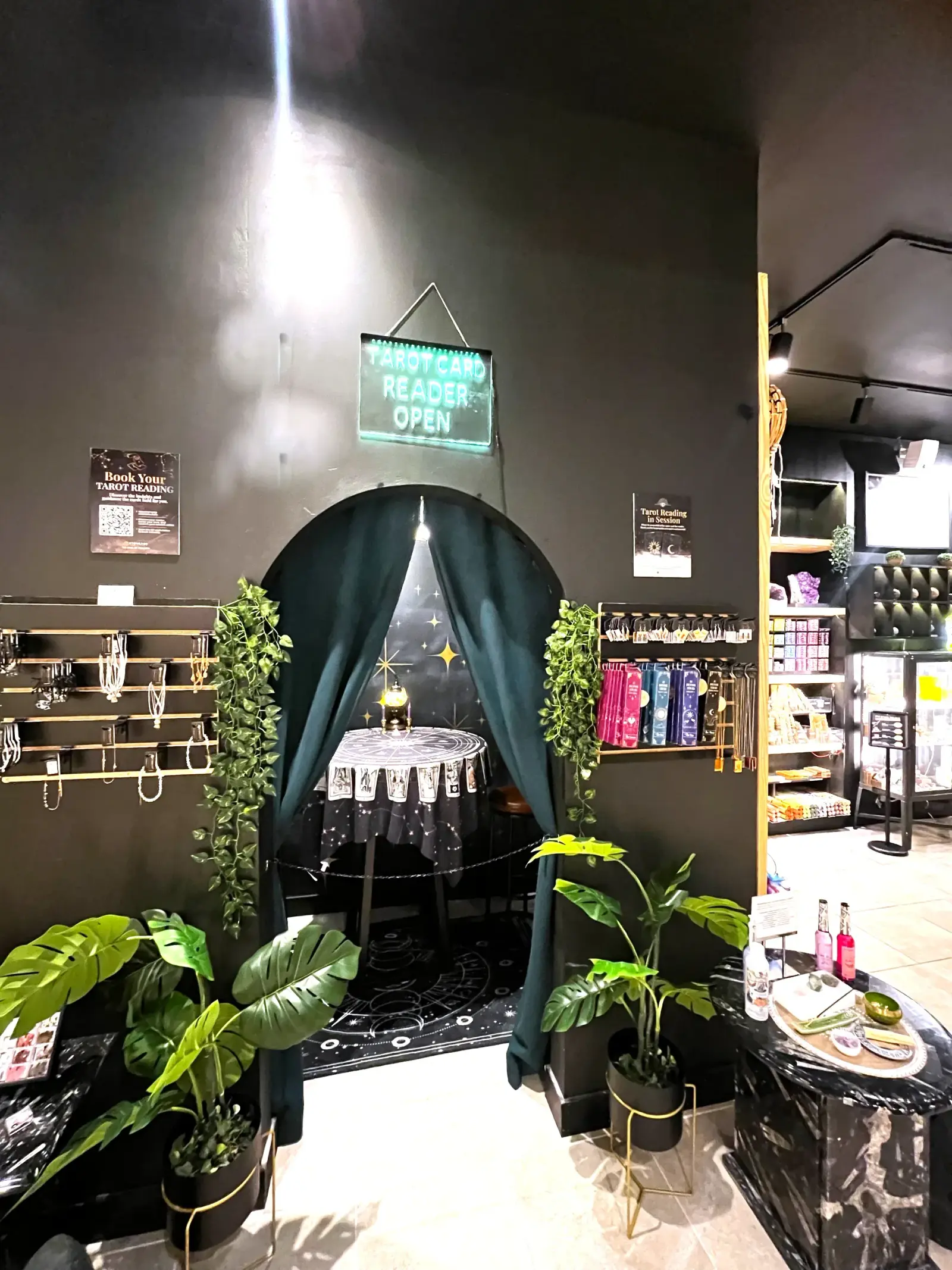 The tarot reading nook: a teal curtain drawn back from an arched alcove, a zodiac cloth on a round table, a lit candle and a neon sign reading Tarot Card Reader Open