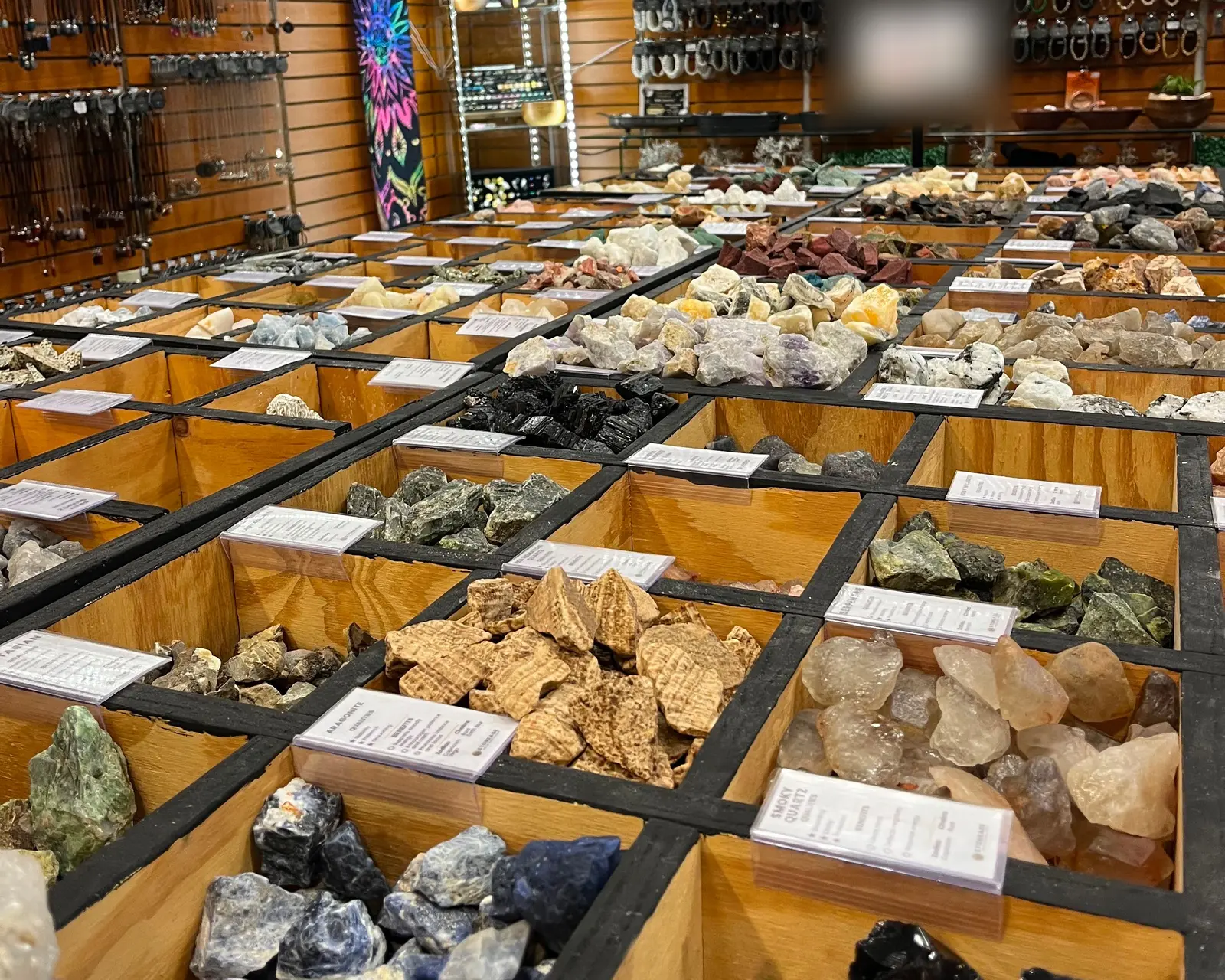 Open timber bins of rough stone at the Cross Creek Mall store, each compartment a different mineral with a written label, walls of hanging jewellery behind