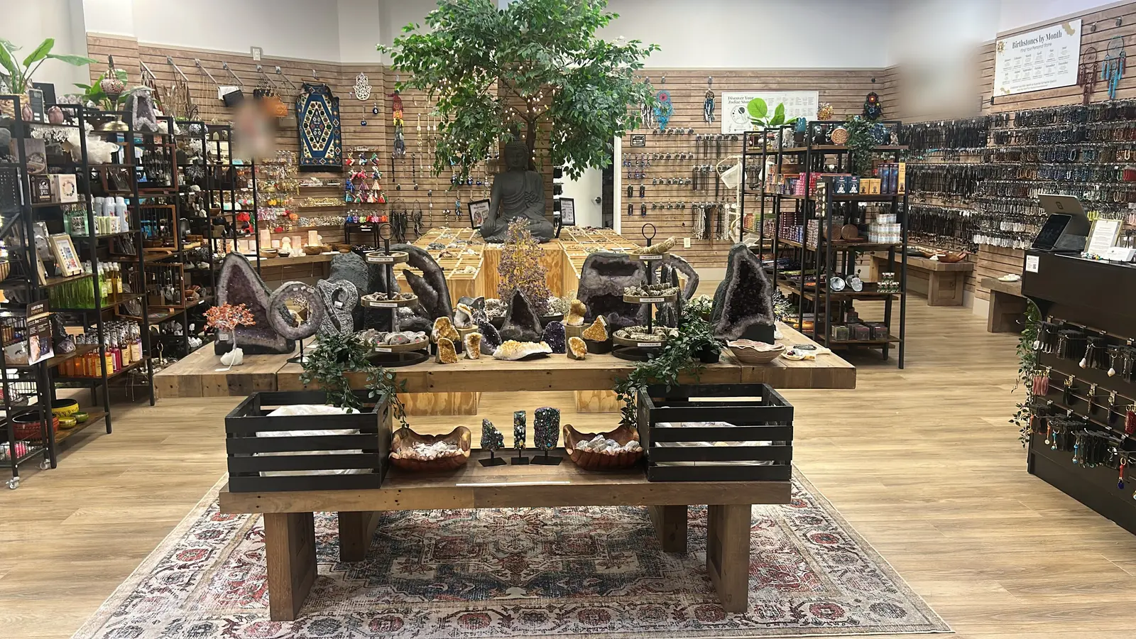 Inside the Concord Mills store: a seated stone figure and a lit tree on the centre line, a timber plinth tiered with amethyst and citrine geodes, crates of tumbled stone on a patterned rug, and walls of hanging jewellery either side