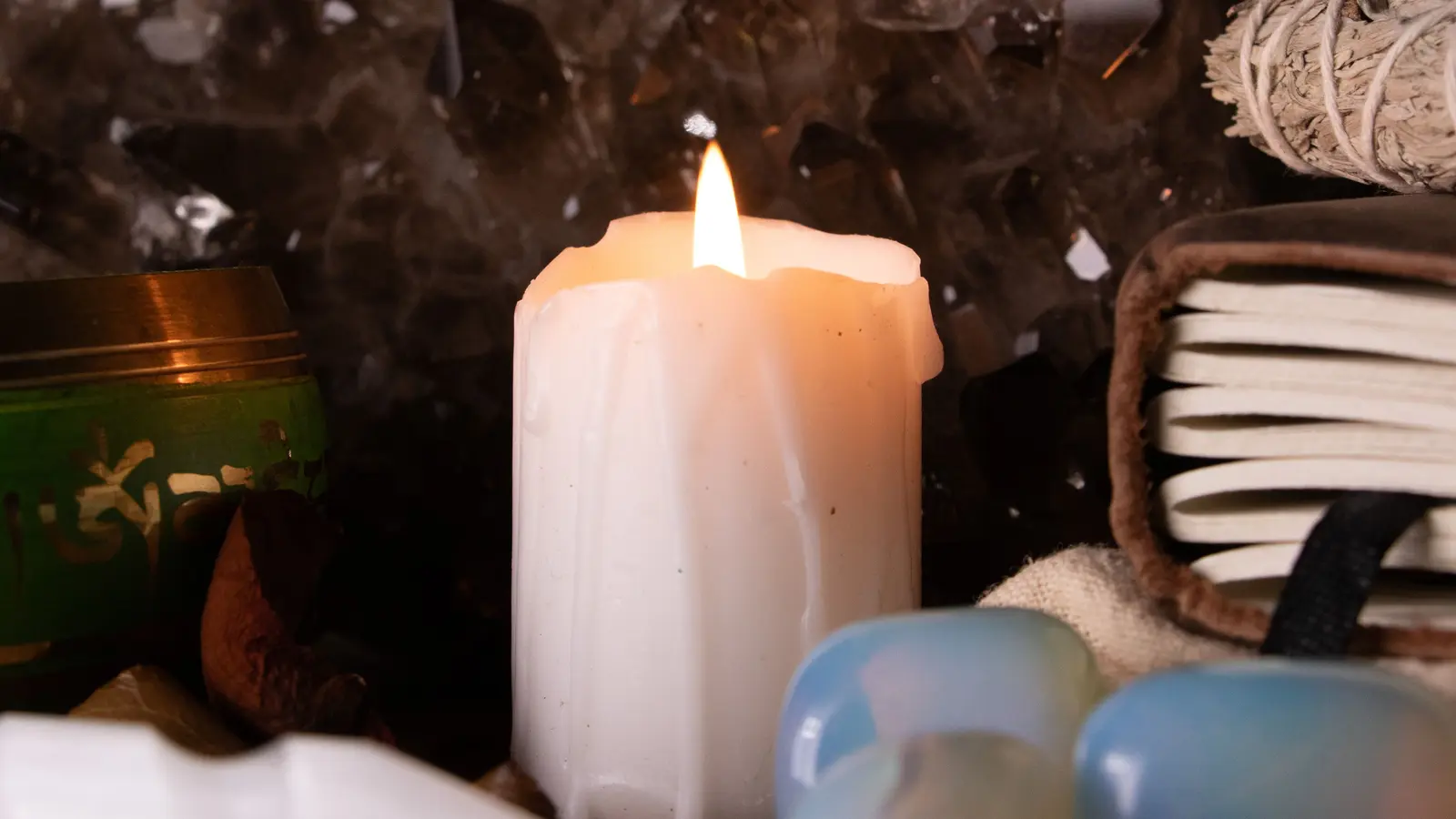 A still life against a large smoky quartz cluster: a lit pillar candle, a green enamelled singing bowl, a leather-bound journal, a bundle of white sage, a white selenite wand and three pale blue-white tumbled pieces on dark timber