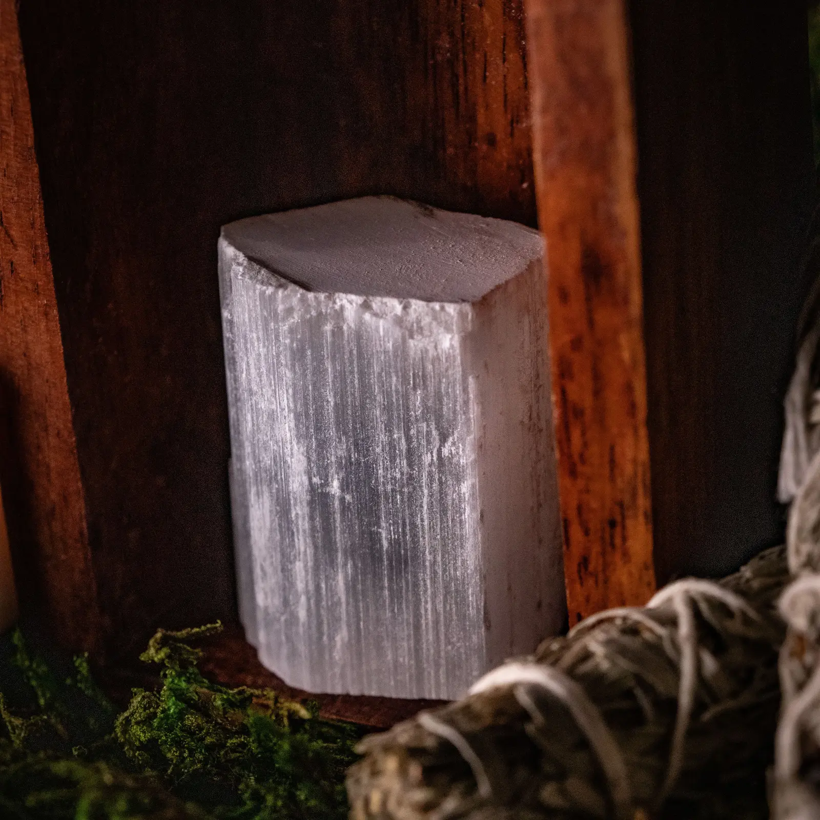 A block of selenite standing on moss, its fibres running the length of the stone, a timber post behind it